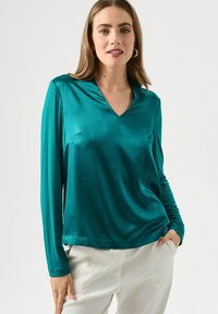 Maglietta a maniche lunghe di colore teal in un tessuto liscio e lucido con scollo a V e vestibilità rilassata; abbinata a pantaloni di colore chiaro.