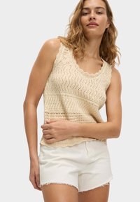 Top de ganchillo beige con escote en forma de ola y patrón de zigzag, combinado con shorts de mezclilla blancos desgastados.