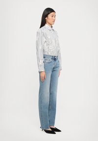 Ριγέ πουκάμισο με λουλουδάτες λεπτομέρειες, ανοιχτόχρωμο τζιν παντελόνι και μαύρα μυτερά παπούτσια. Το μοντέλο στέκεται στο πλάι με φόντο απλό.