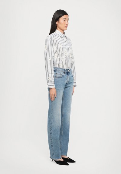 Črtasta srajca s cvetličnimi detajli, svetlo modri kavbojki in črni koničasti čevlji. Model stoji v stranskem profilu pred enostavno ozadjem.