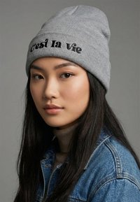 Cappello a maglia grigio con la scritta "C'est la vie" ricamata in nero. Indossato con un dolcevita beige e una giacca di jeans, caratterizzato da una texture a coste.