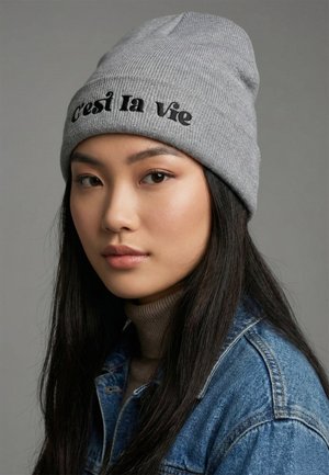 Cappello a maglia grigio con la scritta "C'est la vie" ricamata in nero. Indossato con un dolcevita beige e una giacca di jeans, caratterizzato da una texture a coste.