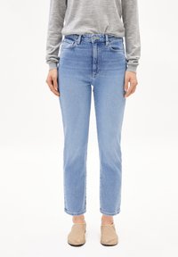 Jean taille haute bleu clair avec une coupe droite, cinq poches et un léger délavage. Associé à une chemise à manches longues grise et des chaussures beiges.