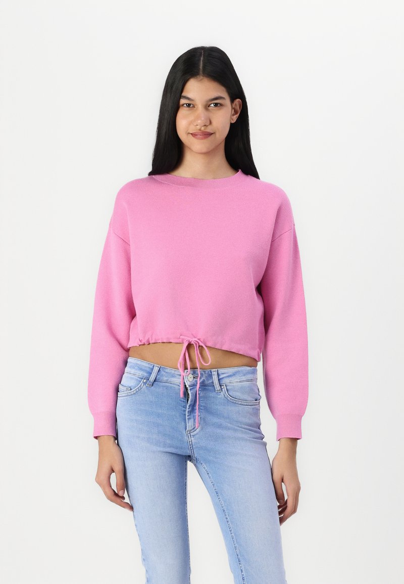 Pull en rose à manches longues et cropped, avec un cordon de serrage à l'ourlet. Porté avec un jean en denim bleu clair, au design et à la texture minimalistes.