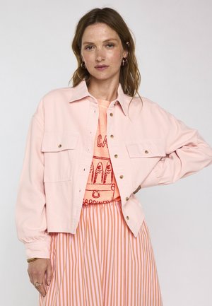 Vrouw met lichtbruin haar die een lichtroze jasje met knopen draagt over een oranje T-shirt met grafische print en een roze-wit gestreepte rok, poserend met hand in de zij.