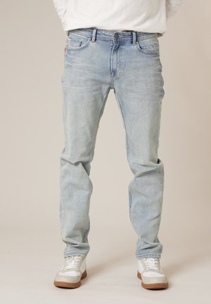 Man wearing light blue jeans and white sneakers standing against plain background.
