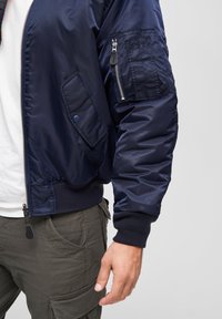 Námořnická modrá saténová bomber bunda s ribbenými manžetami, zipovou kapsou na rukávu a bočními kapsami. Hladký střih s volným designem.