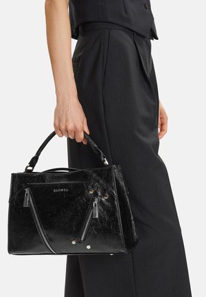 Mano sosteniendo un bolso de cuero negro con textura, con dos bolsillos con cremallera, tachuelas plateadas y asas cortas, vestido con pantalones negros a medida.