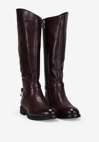 Botas de cuero marrón hasta la rodilla con un acabado suave, cremallera lateral y un detalle de correa con una hebilla metálica. Suela de goma plana y baja.