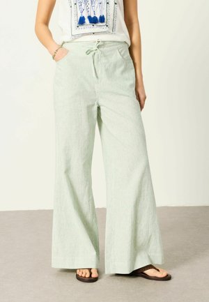Vrouw draagt lichtgroene broek met brede pijpen en trekkoord in de taille, wit mouwloos topje met blauwe kwastdetails, en bruine teenslippers.