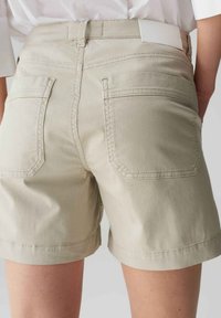 Shorts beiges en tissu de coton, avec une taille ajustée, deux poches arrière et des détails cousus. Texture lisse avec un design décontracté.