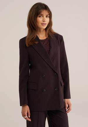 Donna con capelli castani che indossa un blazer doppiopetto a righe sottili scure e pantaloni abbinati su sfondo chiaro.