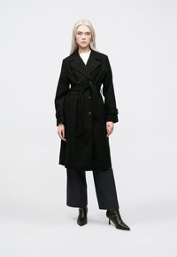 Cappotto nero con un design doppiopetto, vita cinturata e ampi rever. Presenta tasche a pattina e una linea dritta, lunga fino al ginocchio.