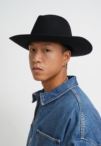 Schwarzer Filzhut mit breiter Krempe, getragen mit einer blauen Jeansjacke, die einen Kragen und einen Reißverschluss hat. Das Subjekt trägt Ohrringe und hat einen neutralen Gesichtsausdruck.