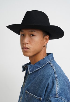 Black felt wide-brimmed hat worn with a blue denim jacket featuring a collar and a zip. Subject has earrings and a neutral expression.