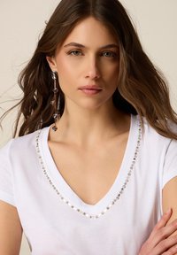Mujer con cabello largo y castaño, vestida con una camiseta blanca de cuello en V con ribete de estrellas plateadas y un pendiente largo colgante, mirando al frente con los brazos cruzados.