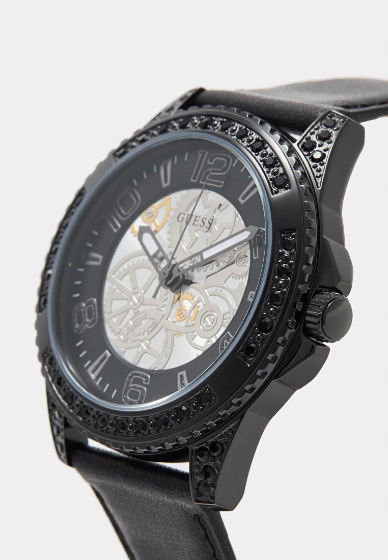 Reloj Guess negro con un dial esqueletón, engranajes plateados visibles, acentos de cristal negro y una correa de cuero negro. Diseño de caja redonda.