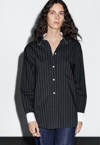 Black striped shirt with white collar and cuffs, long sleeves, front buttons, and a relaxed fit. Paired with navy jeans.