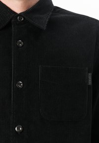 Camisa de pana negra con cierre de botones en la parte delantera, gran collar, un bolsillo en el pecho y una sutil textura acanalada. Botones redondos y detalles de etiqueta visibles.