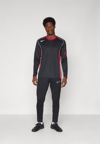 Set sportiv negru format dintr-o bluză cu mânecă lungă cu accente roșii și pantaloni asortați. Logo-ul Nike expus proeminent în alb.