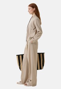 Beige bomberjack met ritszakken, gecombineerd met losse beige broeken. Model houdt een grote zwart-beige gestreepte schoudertas vast.