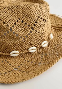 Chapeau en paille tressée marron avec des trous décoratifs et une bande de cinq coquillages blancs autour de la calotte, posé sur une surface claire.