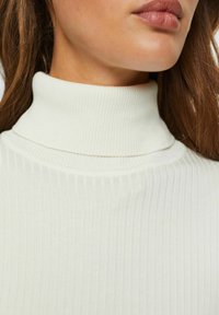 Kvinna i en ribbad vit polotröja, som visar halsen och nedre delen av ansiktet med långt brunt hår.