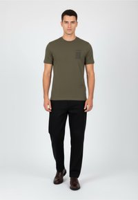 Olivovozelené tričko s krátkým rukávem s texturovanou černou kapsou, spárované s černými kalhotami a hnědými botami. Jednoduché pozadí.