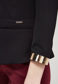 Blazer noir avec une accent en or aux poignets, présentant une plaque logo argentée et un tissu texturé. Comprend des détails subtils au niveau des manches et des poches.