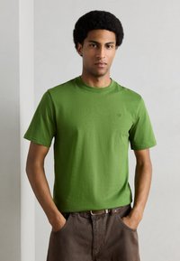 Πράσινο βαμβακερό μπλουζάκι με στρογγυλή λαιμόκοψη και κοντά μανίκια, με διακριτικό λογότυπο στο στήθος. Φορι έται με καφέ παντελόνια.