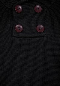 Svart stickad tröja med en texturerad tyg och två stora bourgognefärgade knappar på framsidan som ger detalj åt halsringningen.