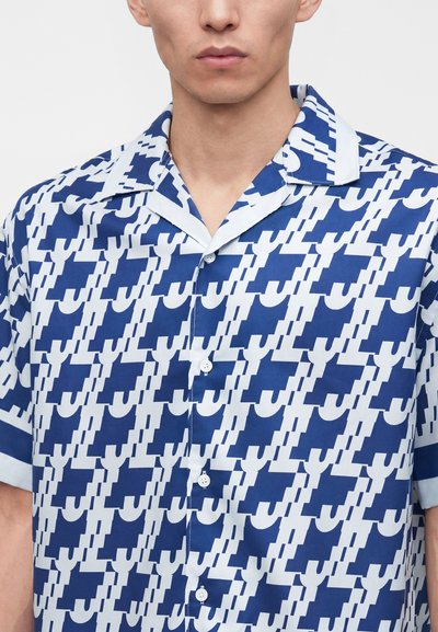 Hombre con camisa de manga corta abotonada con un patrón geométrico abstracto azul y blanco y un cuello abierto.