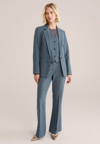 Ušitý oblek tři v jednom z jemné modré látky, zahrnující přiléhavý blazer, vestu se třemi knoflíky a rozšířené kalhoty. Špičaté béžové podpatky.