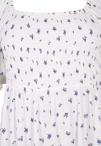 Bílé šaty s malými fialovými a zelenými květinovými vzory, nařasený korzet a splývavá sukně s texturovaným povrchem.