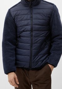 Giacca blu navy con un frontale imbottito e trapuntato e maniche in peluche testurizzate. Presenta tasche con zip e un colletto alto per maggiore calore.