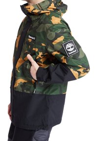 Kamouflagejacka i gröna, svarta och orange tyger. Har en dragkedja framtill, huva, elastiska muddar och en Timberland-logotyp på ärmen.