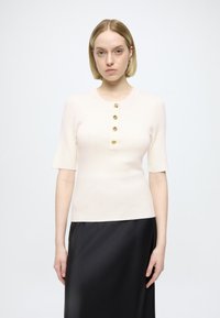 Bordázott krémszínű rövid ujjú felső, amelyet az 5 arany gombdíszítés a nyakkivágásnál díszít, fekete szatén szoknyával párosítva.