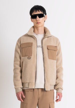 Chaqueta de forro polar beige con dos bolsillos en el pecho marrones, cierre de cremallera y puños de canalé. Lleva puesta encima de un sudadera de color claro y pantalones cargo.