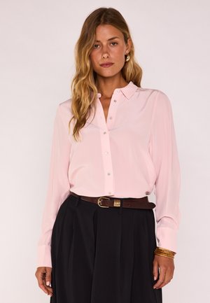 Vrouw met golvend blond haar, gekleed in een lichtroze blouse met knopen, een zwarte rok, een bruine riem, gouden oorringen en gouden armbanden.