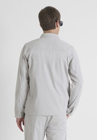 Camisa de botonadura en gris claro, con relieve, de manga larga, corte recto y cuello de punta. Llevada con pantalones a juego, vista desde la parte de atrás.