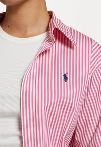 Camicia button-up a righe rosa e bianche con un logo blu, indossata sopra un top a righe bianco; presenta un colletto classico e maniche lunghe.