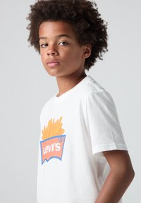 Bílý bavlněný tričko s barevným grafickým potiskem hranolků v boxu s textem "Levi's". Krátké rukávy, kulatý výstřih, ležérní střih.