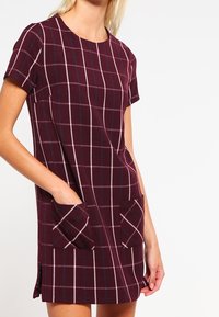 Vestido de manga corta, a cuadros color granate con grandes bolsillos parche, con líneas blancas y azules, hecho de tela texturizada, con aberturas laterales.