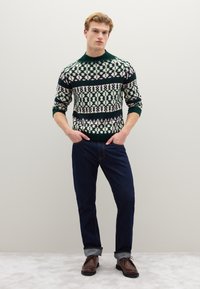 Grün- und cremefarben gemusterter Strickpullover mit Rundhalsausschnitt, kombiniert mit dunkelblauen Jeans und braunen Schnürschuhen. Strukturiertes Strickdetail sichtbar.