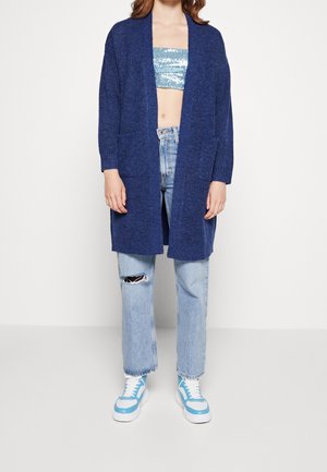 Cardigan lavorato a maglia blu navy con maniche lunghe, apertura frontale e due tasche anteriori, indossato sopra un top corto con paillettes e jeans strappati di un blu chiaro.