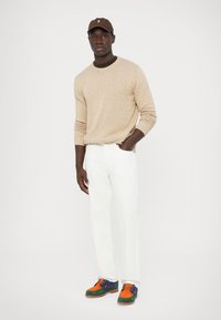 Bézs kötött pulóver, fehér, egyenes szárú nadrág, narancs, kék és zöld díszítésekkel ellátott, többszínű sportcipők és barna sapka.