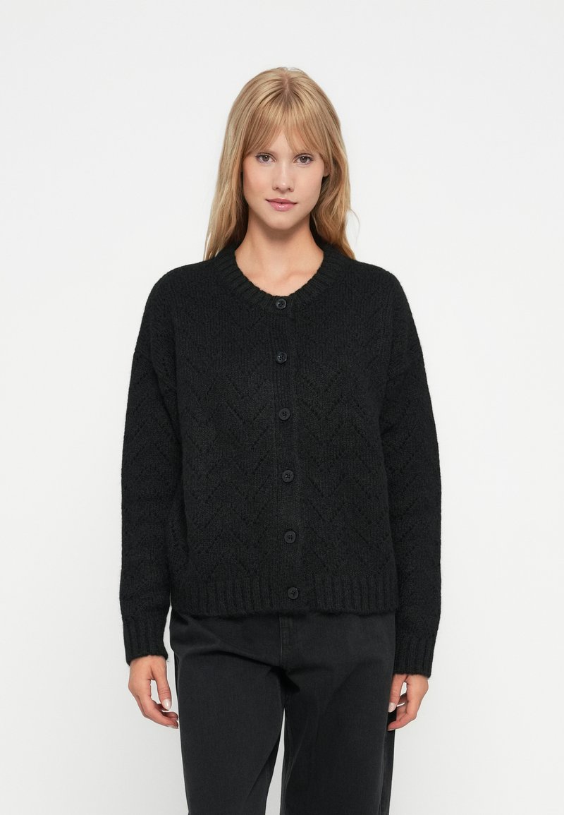 Svart strikket cardigan med chevronmønster, rund hals og seks knapper foran. Har lange ermer og ribbestrikkede mansjetter.