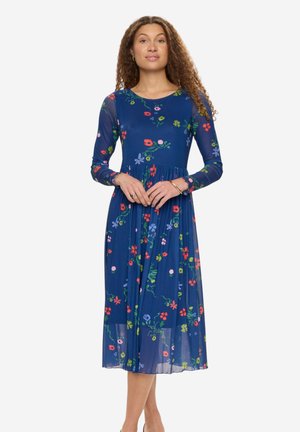 Kobieta o kręconych włosach w granatowej, długorękawowej sukience midi z kolorowymi kwiatowymi wzorami, stojąca z rękami splecionymi przed sobą.