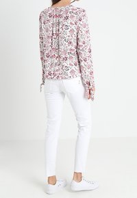 Blütenmuster langärmlige Bluse in Weiß mit rosa und grünen Mustern. Getragen mit weißen, figurbetonten Hosen und lässigen weißen Sneakern.