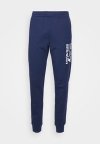 Mörkblå sweatpants i bomullsblandning med elastisk midja, smalnande ben och en vit logotyp tryckt på vänster sida.